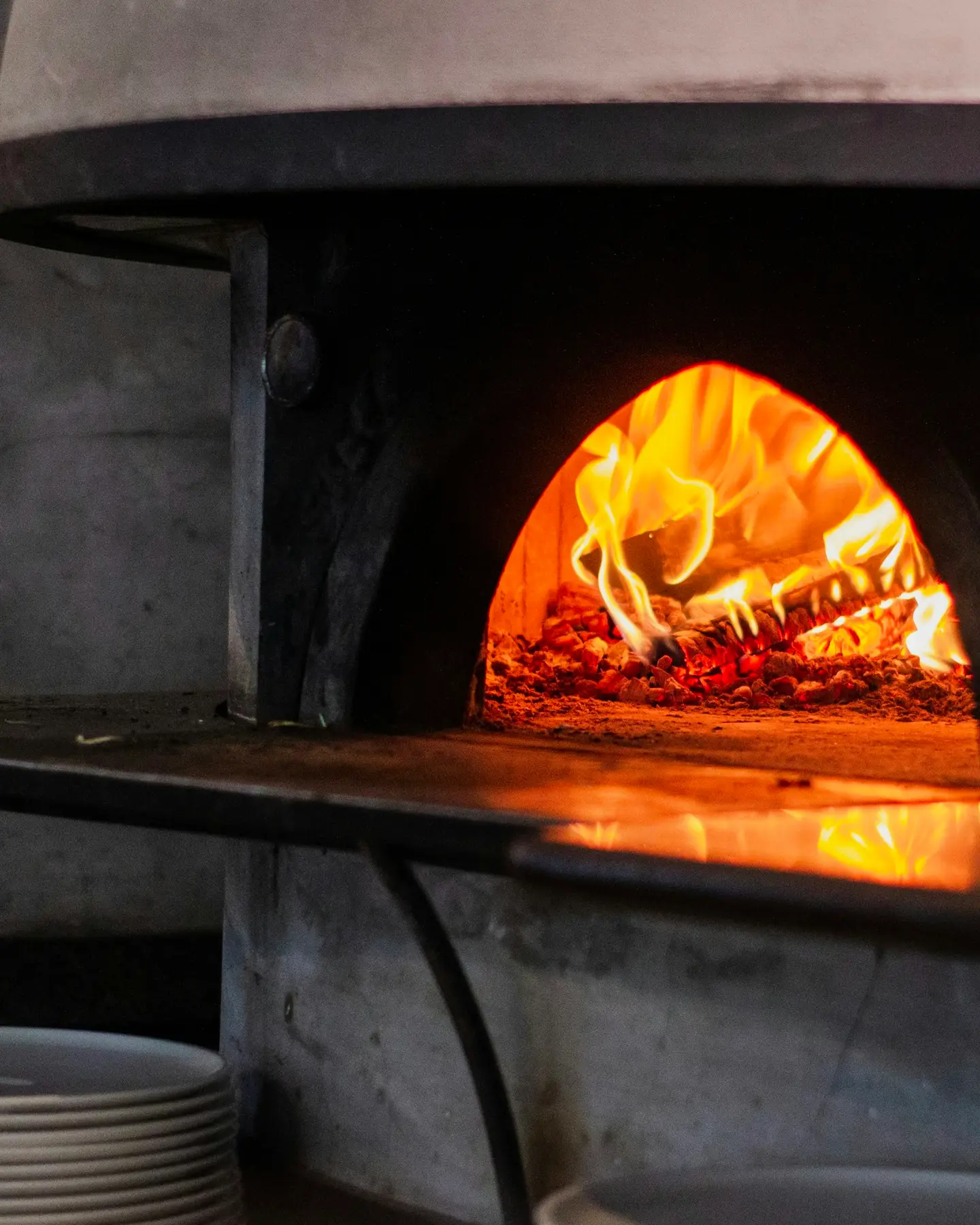 Forno a lenha aceso, com uma pizza lá dentro e pratos empilhados ao lado.