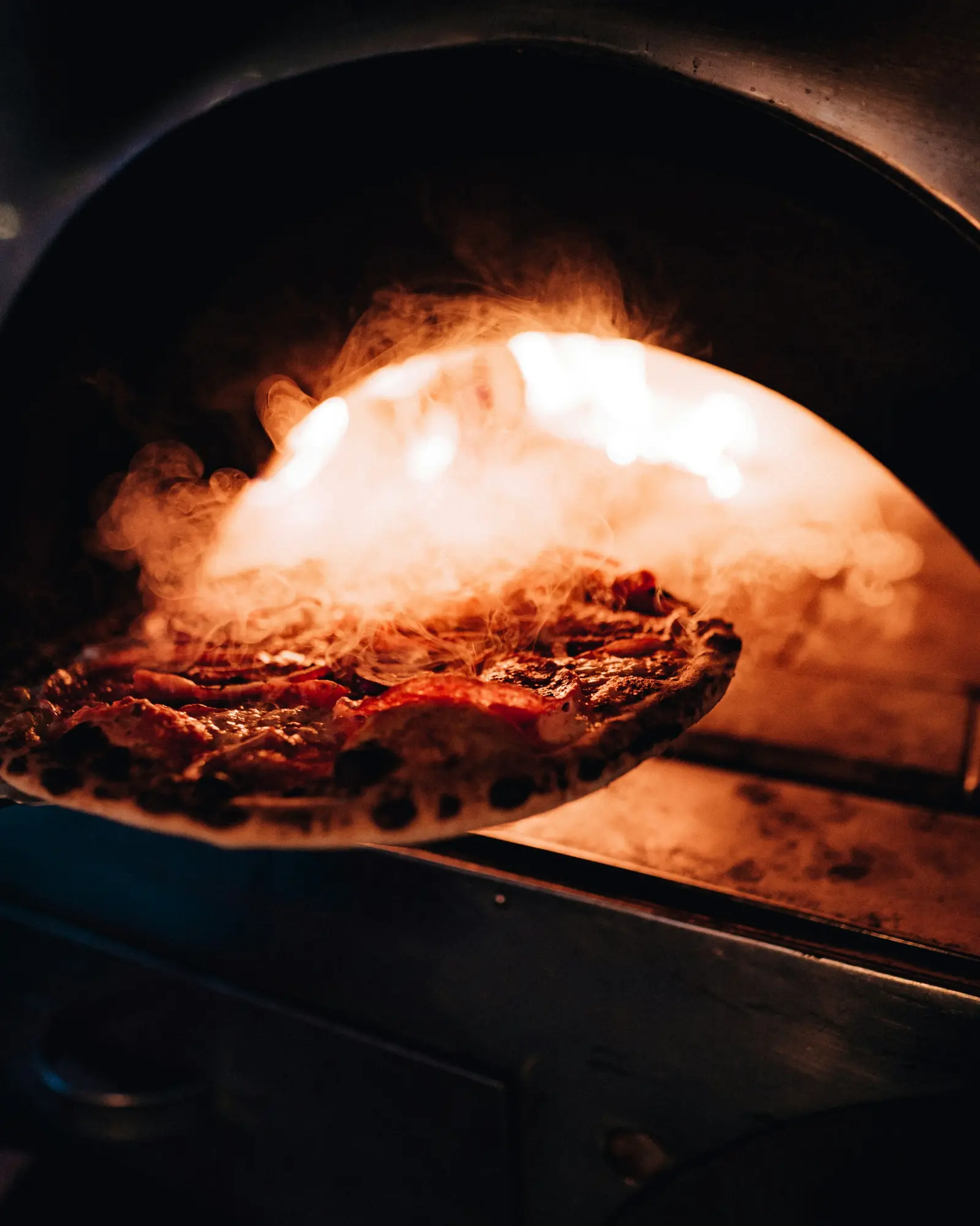 Pizza saindo do forno a lenha numa pá de metal, com o fogo aceso ao fundo.